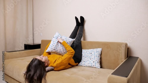 Bored woman lies lifting legs across brown sofa and throws pillows from idleness catching at home slow motion