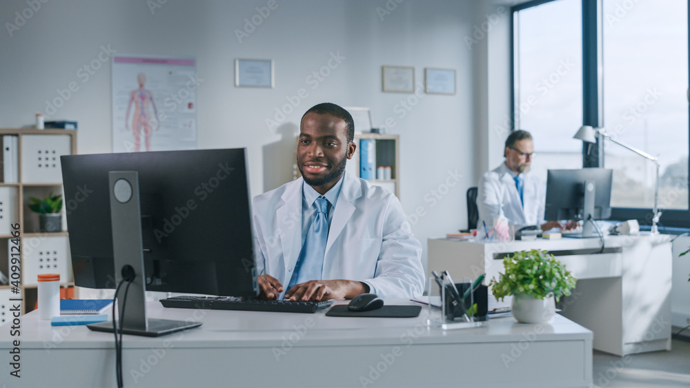 Happy Smiling African American Medical Doctor is Working on a Computer in a Health Clinic. Physician in White Lab Coat is Browsing Medical History Behind a Desk in Hospital Office. 