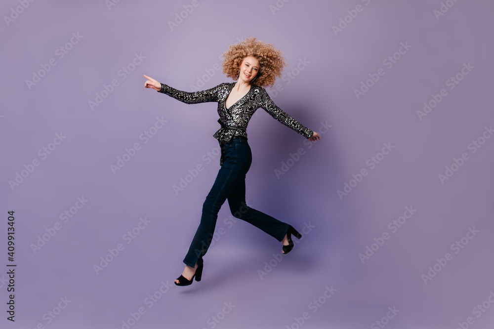 Cheerful slender woman in stylish jeans and disco blouse jumping on lilac background