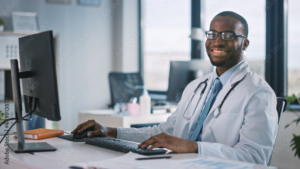 African American Family Medical Doctor in Glasses is Working on a Computer in a Health Clinic. Successful Black Physician in White Lab Coat Looks at the Camera and Smiles in Hospital Office. African American Family Medical Doctor in Glasses is Working on a Computer in a Health Clinic. Successful Black Physician in White Lab Coat Looks at the Camera and Smiles in Hospital Office.