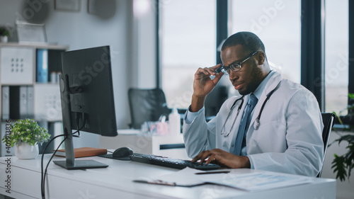 Tired African American Family Medical Doctor in Glasses is Working on a Desktop Computer in a Health Clinic. Physician in White Lab Coat is Browsing Medical History Behind a Desk in Hospital Office. 