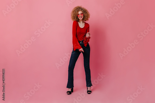 Full-length shot of curly woman in pink glasses posing against isolated background. Girl in red blouse and stylish jeans dancing disco © Look!