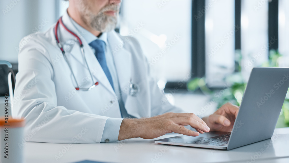 Close Up of a Family Medical Doctor Working on a Laptop Computer in a Health Clinic. Physician in White Lab Coat is Browsing Medical History Behind a Desk in Hospital Office. Close Up of a Family Medical Doctor Working on a Laptop Computer in a Health Clinic. Physician in White Lab Coat is Browsing Medical History Behind a Desk in Hospital Office.