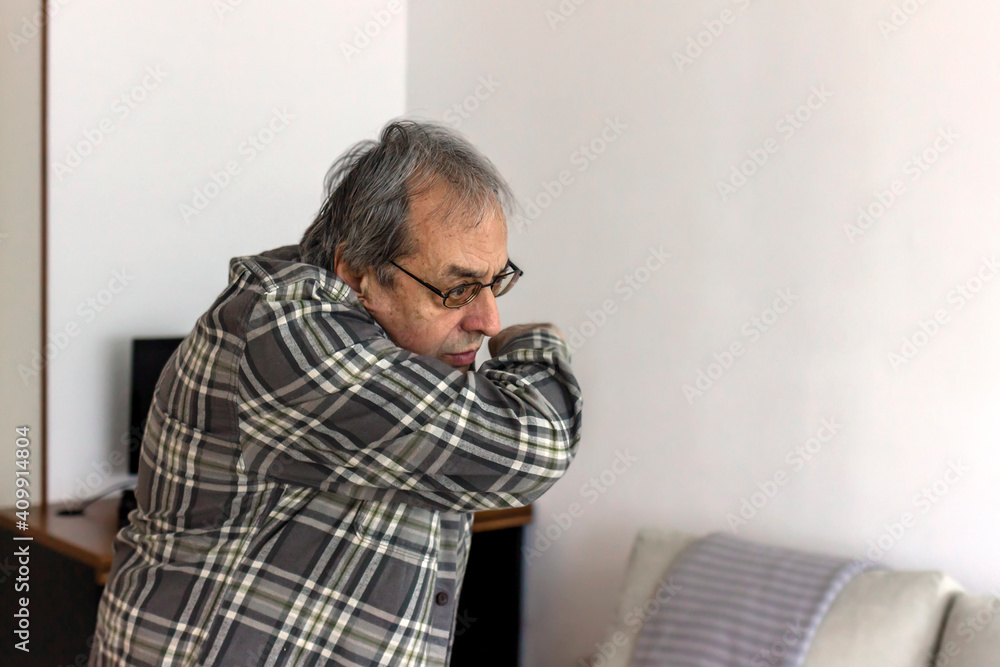 Close up of senior man with eyeglasses coughing into elbow at home. Frustrated elderly man sneezing and coughing into elbow in the living room. Health care and medical concept. Close up. Close up of senior man with eyeglasses coughing into elbow at home. Frustrated elderly man sneezing and coughing into elbow in the living room. Health care and medical concept. Close up.