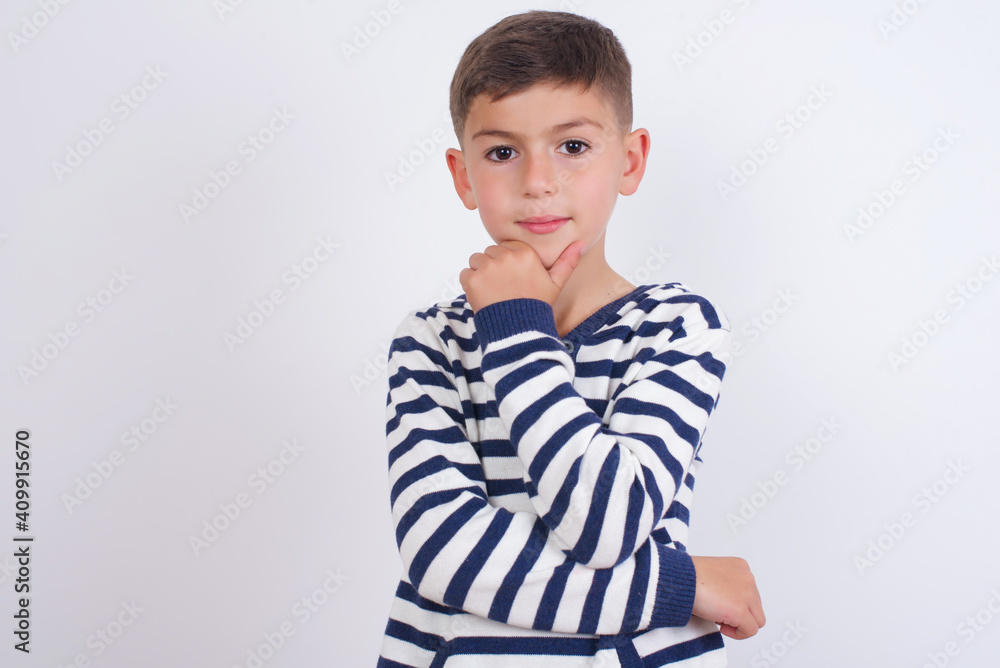 Thoughtful smiling little cute boy kid wearing stripped t-shirt against white wall keeps hand under chin, looks directly at camera, listens something with interest. Youth concept. Thoughtful smiling little cute boy kid wearing stripped t-shirt against white wall keeps hand under chin, looks directly at camera, listens something with interest. Youth concept.