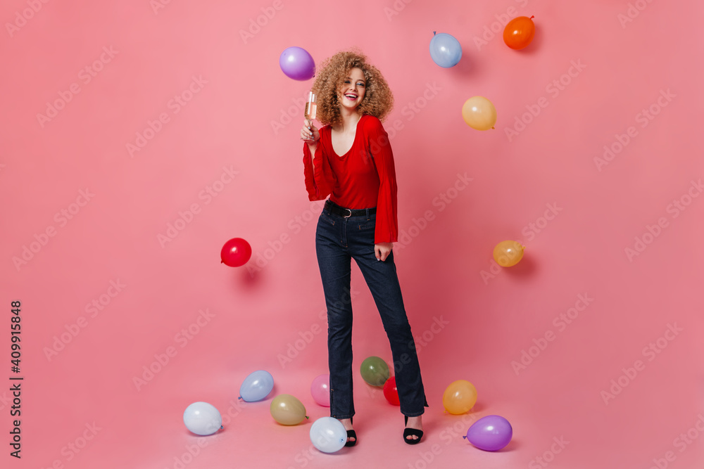 Fototapeta premium Cheerful girl with glass of champagne posing on pink background among balloons. Snapshot of curly woman in red top and denim pants
