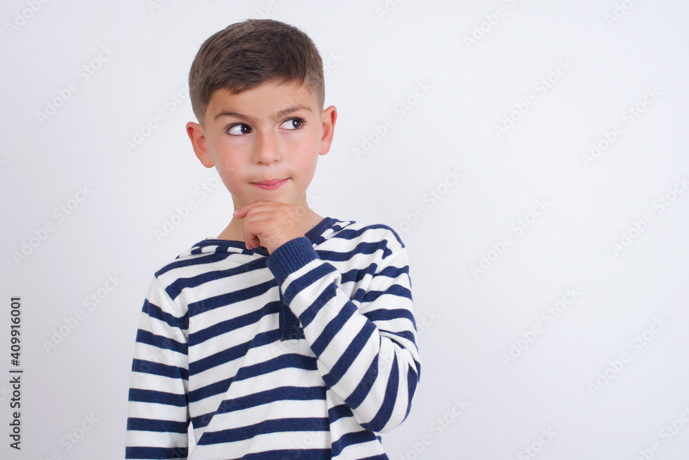 Shot of contemplative thoughtful little cute boy kid wearing stripped t-shirt against white wall keeps hand under chin, looks thoughtfully upwards. Shot of contemplative thoughtful little cute boy kid wearing stripped t-shirt against white wall keeps hand under chin, looks thoughtfully upwards.