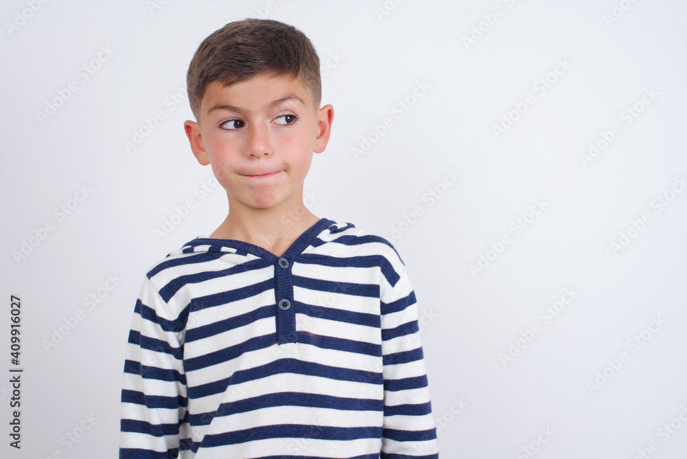 Amazed puzzled little cute boy kid wearing stripped t-shirt against white wall, curves lips and has worried look, sees something awful in front. Amazed puzzled little cute boy kid wearing stripped t-shirt against white wall, curves lips and has worried look, sees something awful in front.