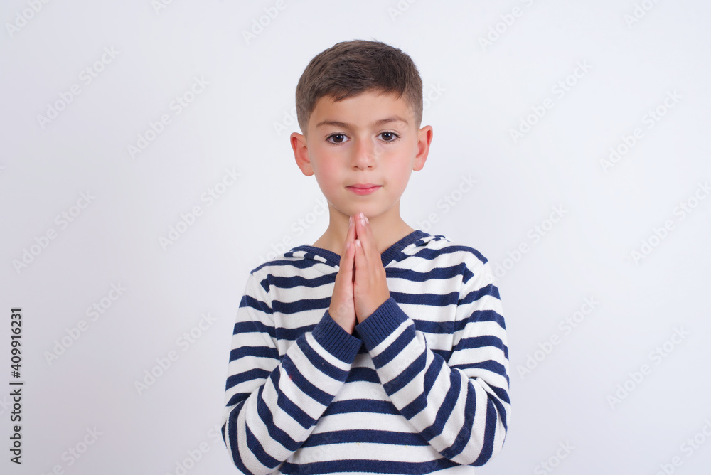 little cute boy kid wearing stripped t-shirt against white wall keeps palms pressed together in front of her having regretful look, asking for forgiveness. Forgive me please. little cute boy kid wearing stripped t-shirt against white wall keeps palms pressed together in front of her having regretful look, asking for forgiveness. Forgive me please.