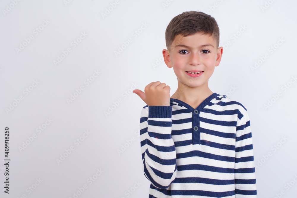 Stupefied little cute boy kid wearing stripped t-shirt against white with surprised expression, opens eyes and mouth widely, points aside with thumb, shows something strange. Advertisement concept. Stupefied little cute boy kid wearing stripped t-shirt against white with surprised expression, opens eyes and mouth widely, points aside with thumb, shows something strange. Advertisement concept.