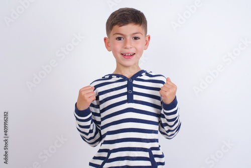 little cute boy kid wearing stripped t-shirt against white wall rejoicing success and victory clenching his fists with joy being happy to achieve her aim and goals. Positive emotions, feelings.