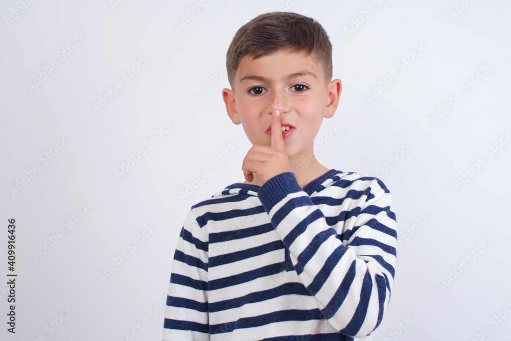little cute boy kid wearing stripped t-shirt against white wall makes hush gesture, asks be quiet. Don't tell my secret or not speak too loud, please! little cute boy kid wearing stripped t-shirt against white wall makes hush gesture, asks be quiet. Don't tell my secret or not speak too loud, please!