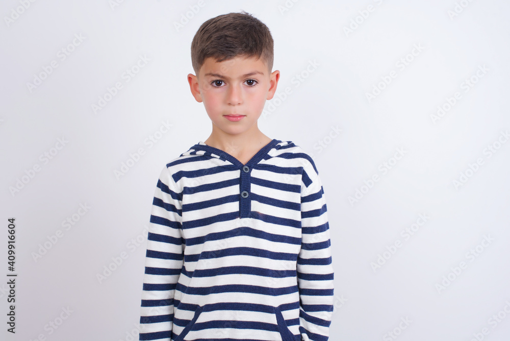 Joyful little cute boy kid wearing red stripped t-shirt against white wall looking to the camera, thinking about something. Both arms down, neutral facial expression. Joyful little cute boy kid wearing red stripped t-shirt against white wall looking to the camera, thinking about something. Both arms down, neutral facial expression.