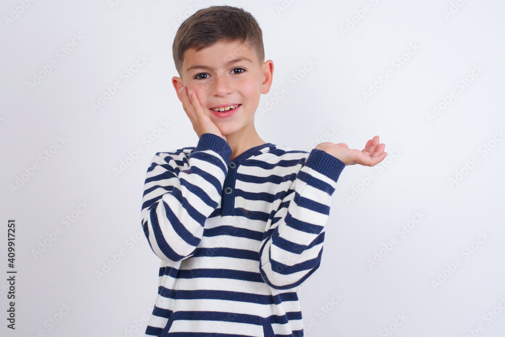 Positive glad little cute boy kid wearing red stripped t-shirt against white wall says: wow how exciting it is, indicates something. One hand on his head and pointing with other hand. Positive glad little cute boy kid wearing red stripped t-shirt against white wall says: wow how exciting it is, indicates something. One hand on his head and pointing with other hand.