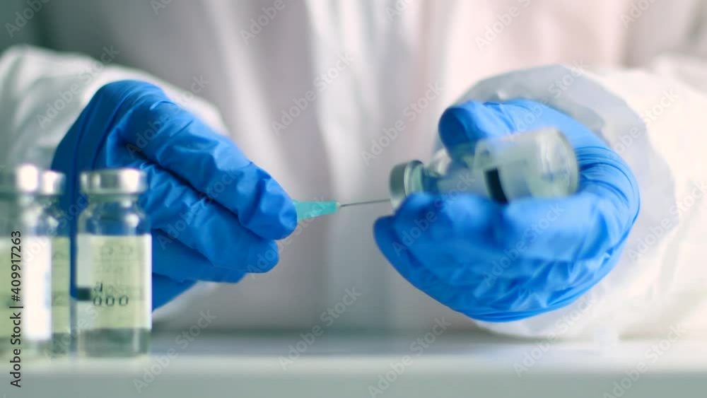 Doctor or nurse hands in medical protective gloves holds syringe and picks up vaccine from a glass vial for an injection against coronavirus, measles or flu disease Doctor or nurse hands in medical protective gloves holds syringe and picks up vaccine from a glass vial for an injection against coronavirus, measles or flu disease