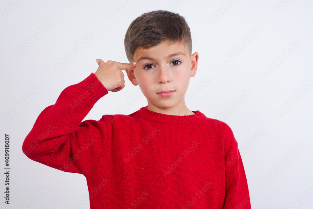 Unhappy Little cute boy kid wearing red knitted sweater against white wall curves lips and makes suicide gesture, shoots in temple with hand, tries kill himself. Unhappy Little cute boy kid wearing red knitted sweater against white wall curves lips and makes suicide gesture, shoots in temple with hand, tries kill himself.