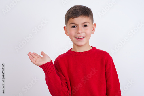 Positive glad Little cute boy kid wearing red knitted sweater against white says: wow how exciting it is, has amazed expression, shows something on blank space with open hand. Advertisement concept.