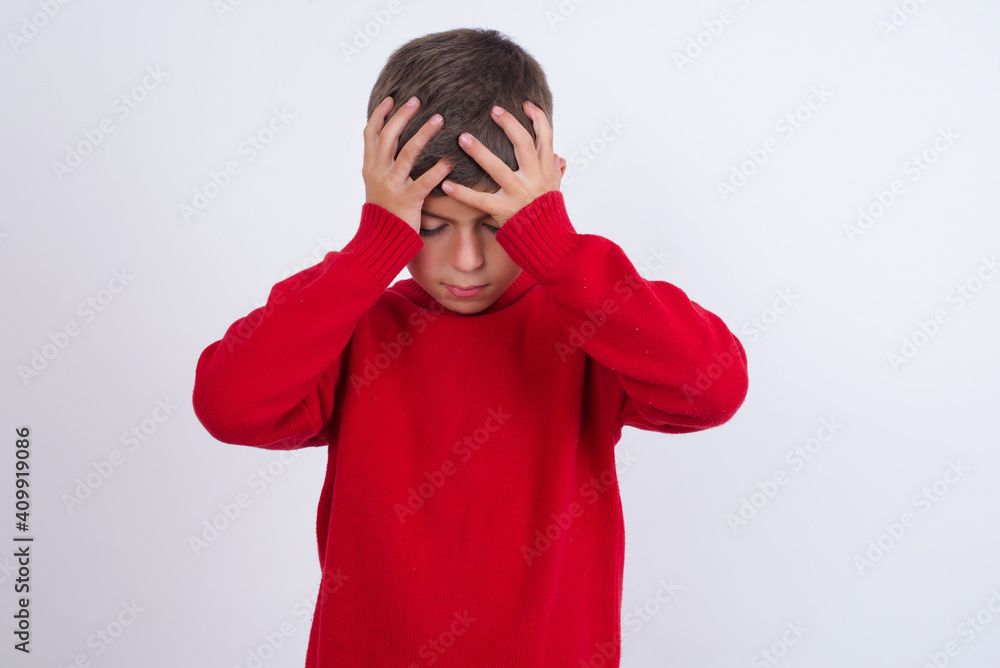 Little cute boy kid wearing red knitted sweater against white wall holding head with hands, suffering from severe headache, pressing fingers to temples Little cute boy kid wearing red knitted sweater against white wall holding head with hands, suffering from severe headache, pressing fingers to temples