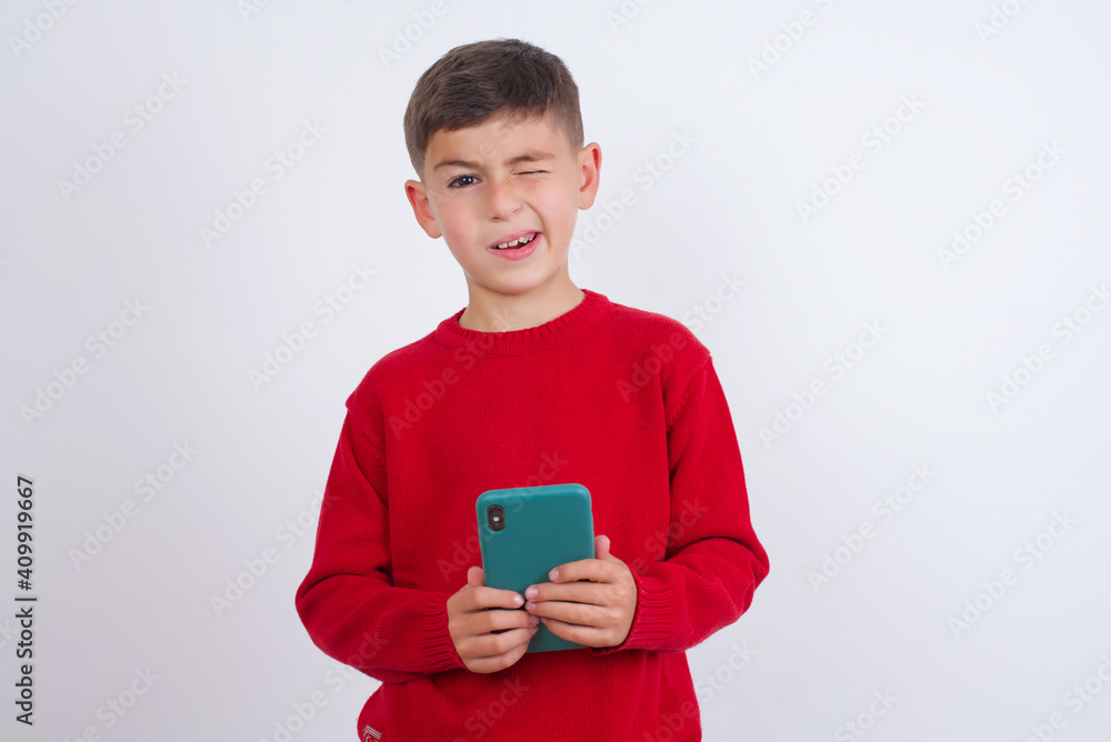 Pleased Little cute boy kid wearing red knitted sweater against white wall using self phone and looking and winking at the camera. Flirt and coquettish concept. Pleased Little cute boy kid wearing red knitted sweater against white wall using self phone and looking and winking at the camera. Flirt and coquettish concept.