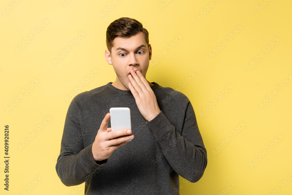 Amazed man covering mouth while holding smartphone on yellow background Amazed man covering mouth while holding smartphone on yellow background