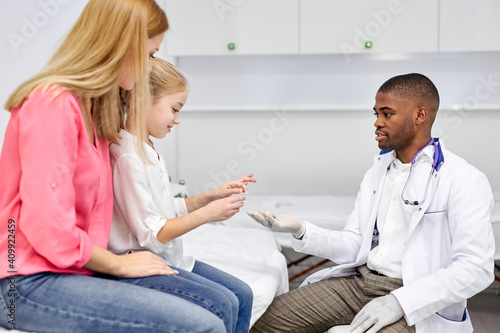 black male doctor have a nice talk with kid girl, showing colourful modern capsules to her, he disposes, work with children in hospital