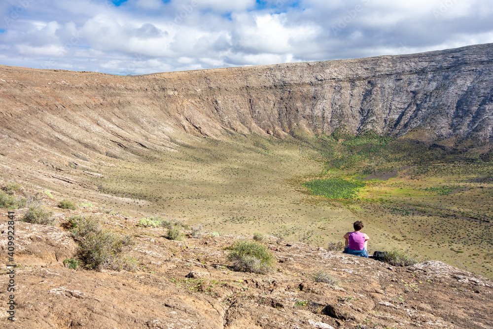 Panoramic view of Caldera Blanca, Timanfaya, Lanzarote,  with a female hiker in a pink shirt sitting on the edge of the crater