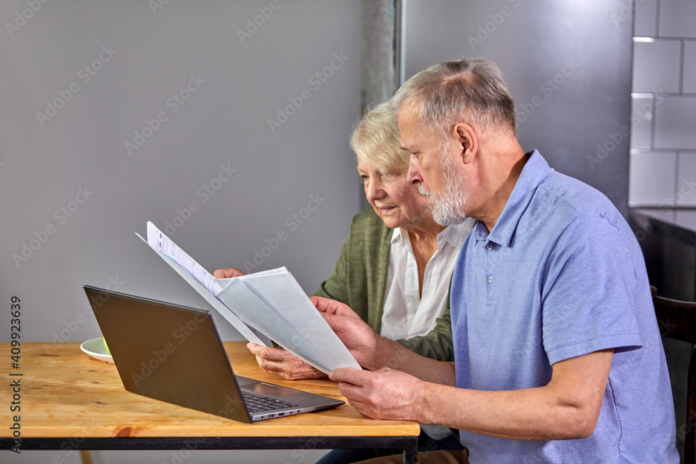 aged couple checking finances at home using laptop, discussing planning budget together, using online banking services and calculator, holding documents in the kitchen aged couple checking finances at home using laptop, discussing planning budget together, using online banking services and calculator, holding documents in the kitchen