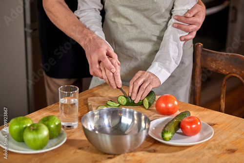 elderly woman cutting a cucumber for a salad in a modern, eco kitchen, preparing food with her husband hugging her from back. at home. cropped people