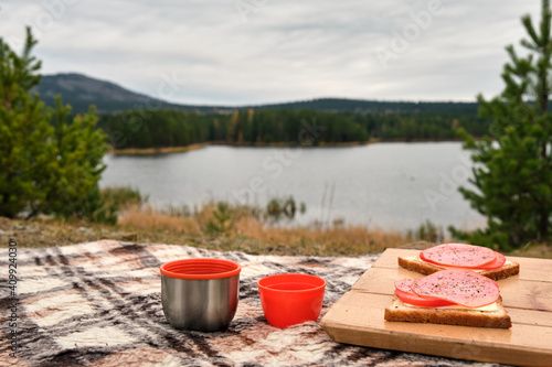 Sandwiches on the wooden stand with orange mugs from thermos on the plaid. Cozy picnic on fresh air. Mountains are visible in the distance.