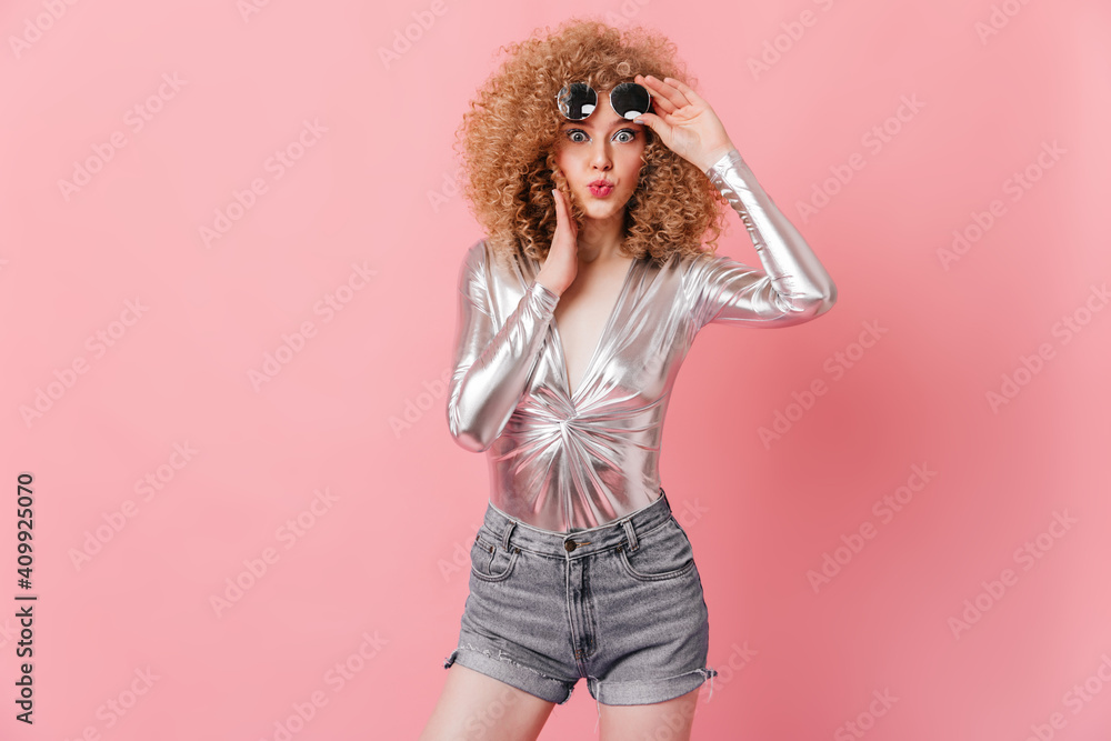 Portrait of charming curly blonde with blue eyes taking off her sunglasses. Woman in shiny top and shorts looks at camera in surprise on pink background Portrait of charming curly blonde with blue eyes taking off her sunglasses. Woman in shiny top and shorts looks at camera in surprise on pink background