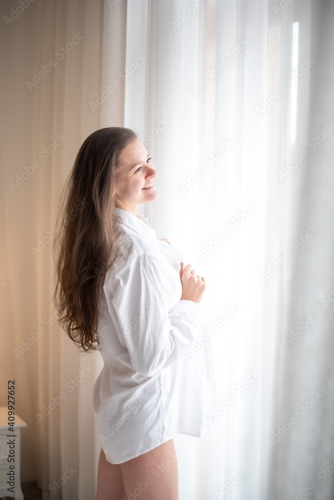 Brunette in mens shirt. Rear view of young woman looking through the window in the morning Brunette in mens shirt. Rear view of young woman looking through the window in the morning
