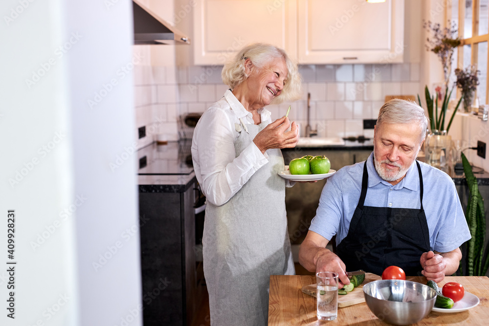elderly couple in the kitchen at home, senior man in apron sits carving fresh vegetables, gray-haired woman holding apples in hands and talking with him elderly couple in the kitchen at home, senior man in apron sits carving fresh vegetables, gray-haired woman holding apples in hands and talking with him