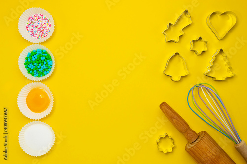 Ingredients, sugar decoration, kitchen tools for baking on the yellow background. Cookie cutters, a rolling pin, an egg whisk. Yolk, sugar, baking decoration in muffin forms. Copy space. Top view.