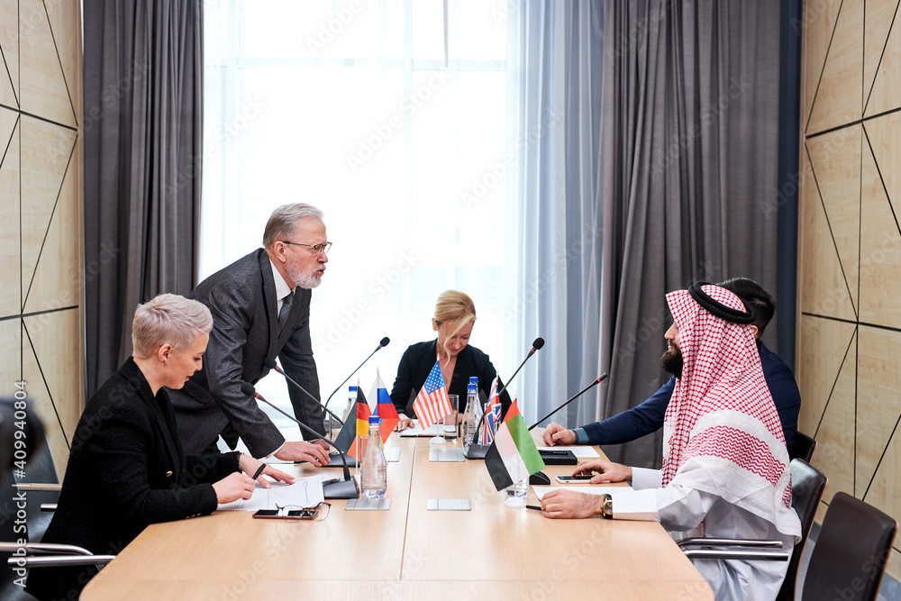 Fototapeta premium senior politician offers his plan of action and share opinion, elderly man in suit talking to multi-ethnic group of partners sitting at desk in boardroom, discussing