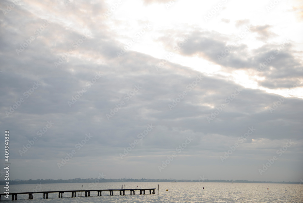 Lake at sunset with clouds and wooden jetty. Sailboats in the waves.