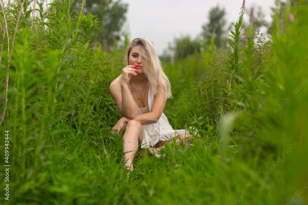 Sexy young blonde girl with red lipstick and summer dress sits among the high green grass in clear summer weather. High quality photo