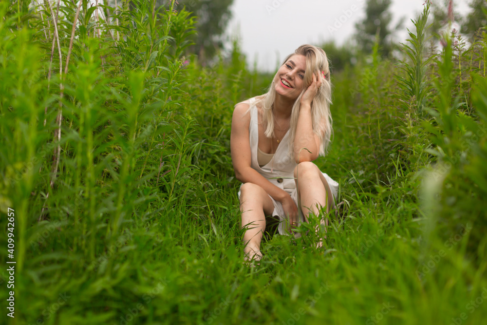 Bright blonde girl in a summery white dress sits among the tall grass in clear summer weather. Summer landscape. Walk out of town. High quality photo