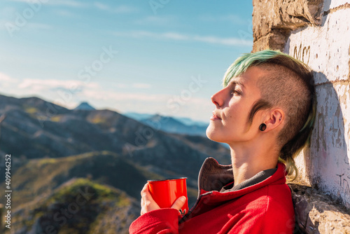 Side view of androgynous traveler standing in highlands on sunny day and enjoying hot drink during trip in El Mazuco with eyes closed photo