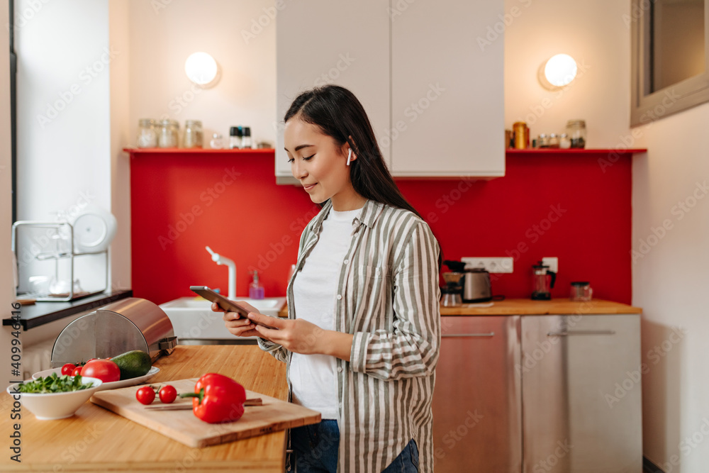 Positive woman in headphones smiles, reads recipe and prepares dinner in kitchen Positive woman in headphones smiles, reads recipe and prepares dinner in kitchen