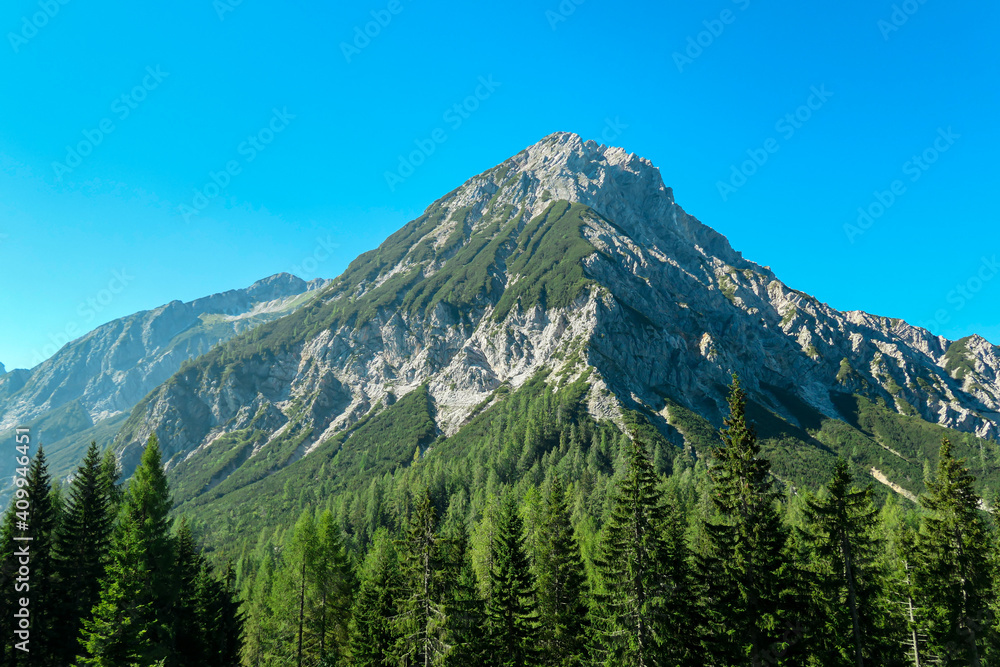 A clear view on Mittagskogel in Austrian Alps. The mountain stands independently. The high mountain is overgrown with a lush forest, with barren and sharp peak. Clear and sunny day. Alpine landscape