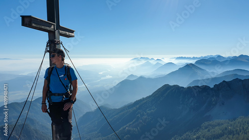 A man with big backpack standing under a metal cross with a panoramic view on the Alps from the top of Mittagskogel in Austrian Alps. Clear and sunny day. A bit of haze in the valley. Outdoor activity