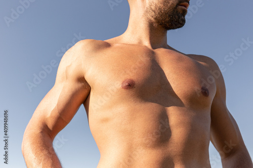 Determined male athlete with strong naked torso standing on grassy hill on sunny day in summer and looking away