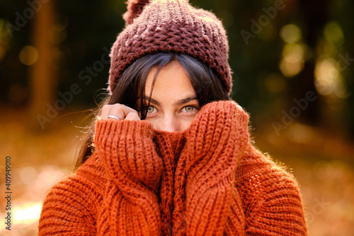 Young female in knitted hat covering face with warm sweater while standing in forest in cold autumn day photo