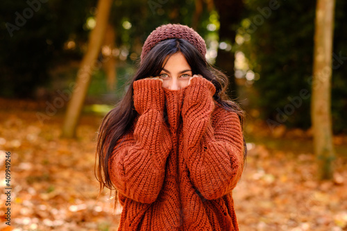 Young female in knitted hat covering face with warm sweater while standing in forest in cold autumn day