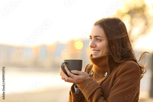 Happy woman drinks coffee in winter looking away on the beach