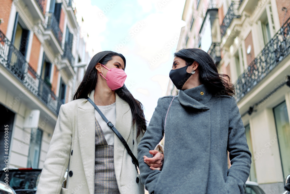 Two happy women talking and walking in the street with their masks on.