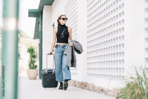 Stylish female tourist with suitcase walking along street in city and looking away photo