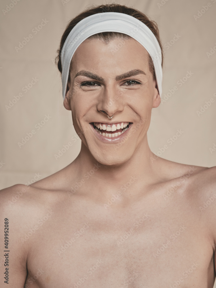 Happy young androgynous male model with makeup and headband looking at camera and laughing against beige background Happy young androgynous male model with makeup and headband looking at camera and laughing against beige background