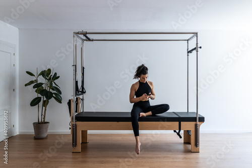 Fit female athlete sitting on pilates cadillac reformer and browsing cellphone while having break during training