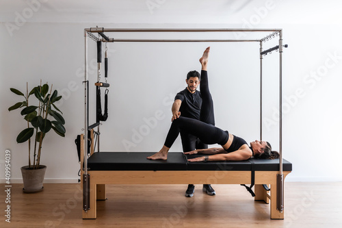 Slim female in sportswear doing exercises on cadillac reformer with help of professional trainer during pilates workout